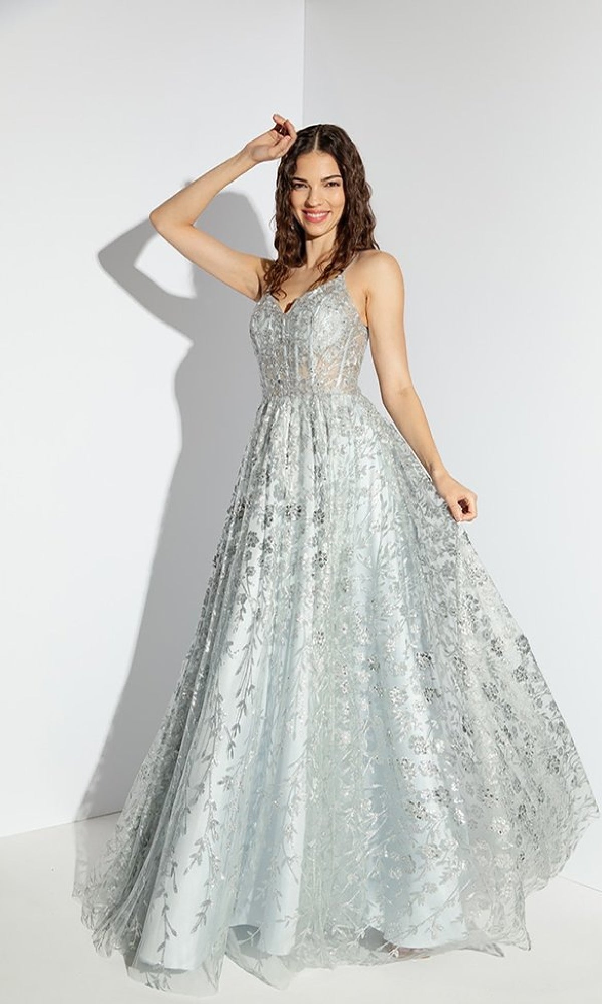 A woman wearing the Long Glitter A-Line Prom Dress: Eureka 9908 in light blue with silver floral embroidery poses against a white background, holding her gown and smiling.